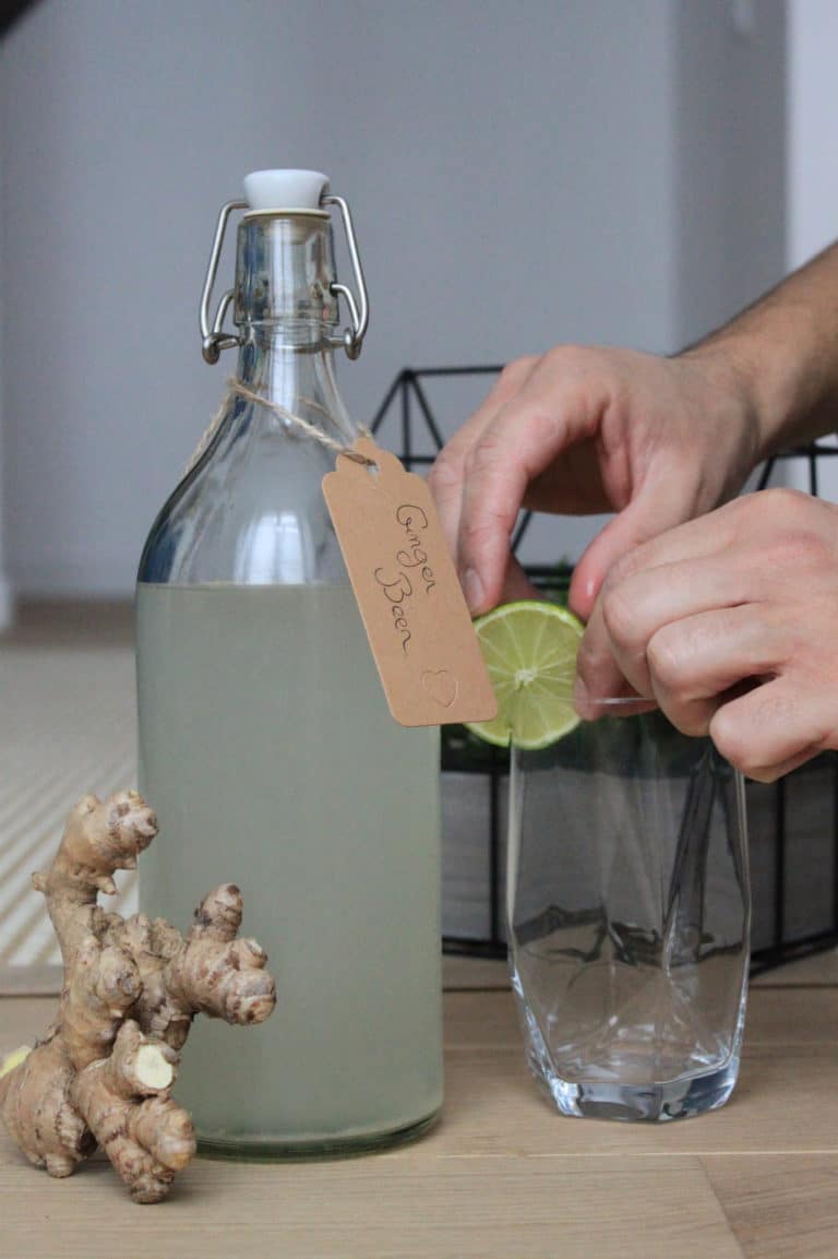 Une bouteille de ginger beer, du gingembre, et des mains qui mettent une rondelle de citron vert sur un verre