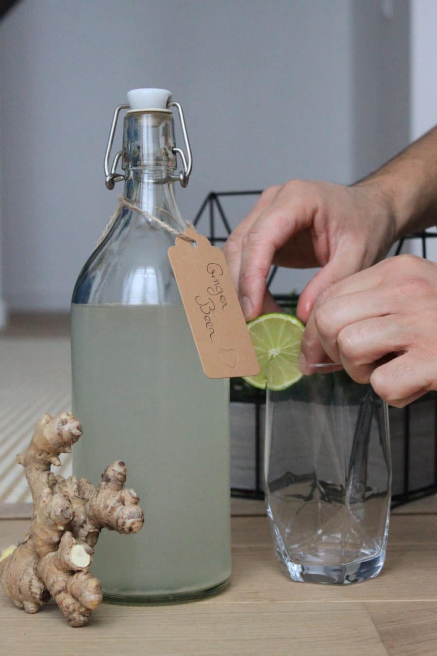 Une bouteille de ginger beer, du gingembre, et des mains qui mettent une rondelle de citron vert sur un verre