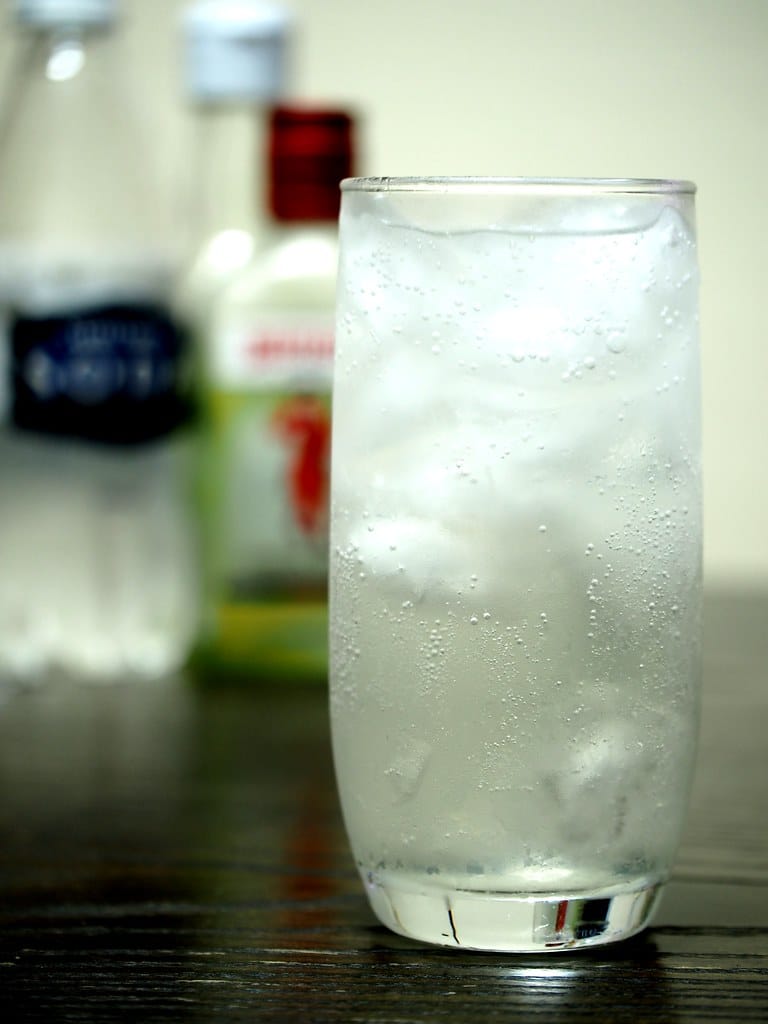 Un grand verre de Tom Collins avec une bouteille de soda water et de gin en arrière plan